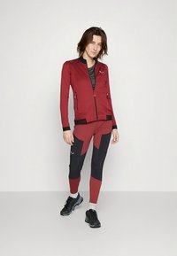 Punane lukuga sportjakk tekstuurse kangaga, koos punaste ja laivaste leggingsidega, millel on värviplokkide disain. Mustad tossud täiendavad välimust.