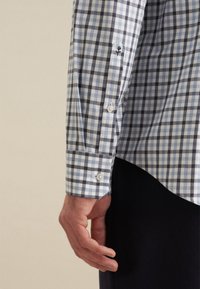 Camicia a quadri blu e bianca con maniche lunghe, polsini con bottoni e dettagli ricamati sui polsi. Il tessuto appare liscio e leggero.
