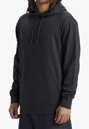 Mand iført en ensfarvet sort pullover-hoodie med frontlomme og snører, parret med mørkeblå bukser.