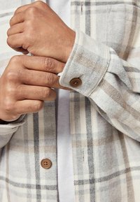 Flanellhemd in beige und grauem Karomuster, mit einem Knopfmanschette und weicher Textur. Braunes Knopfdetail nahe dem Handgelenk verbessert das Design.
