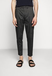 Man som bär mörkgrå slim-fit byxor och svarta slidesandaler, stående mot en enkel ljus bakgrund.