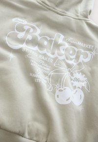 Világos bézs kapucnis pulóver fehér grafikus szöveggel és illusztrációkkal, többek között cseresznyékkel és díszítésekkel, puha, texturált anyagból.