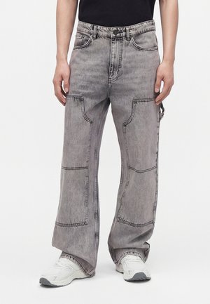 Mann trägt lockere hellgraue Jeanshose mit großen aufgesetzten Taschen und weißen Sneakers vor weißem Hintergrund.