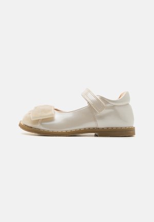 Chaussures Mary Jane pour enfants en matériau crème brillant avec un grand nœud sur le devant, une bride Velcro et une semelle en caoutchouc brun clair.