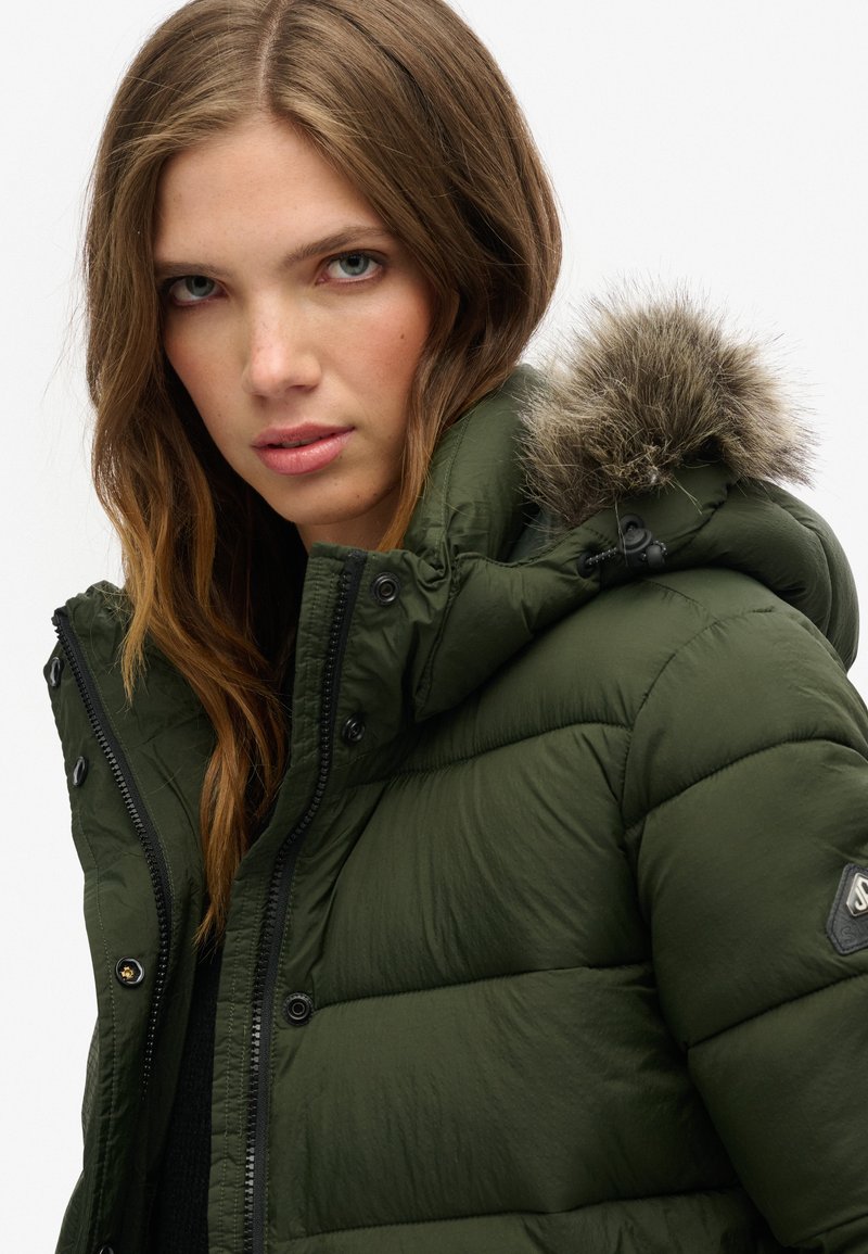 Giacca a vento verde scuro con finitura testurizzata, cappuccio attaccato con bordo in pelliccia sintetica e cerniere e bottoni neri come accessori.