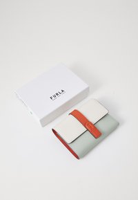 Furla portemonnee met een textured witte leren flap, een muntgroene body en een oranje strap. Geplaatst naast een witte doos met logo.