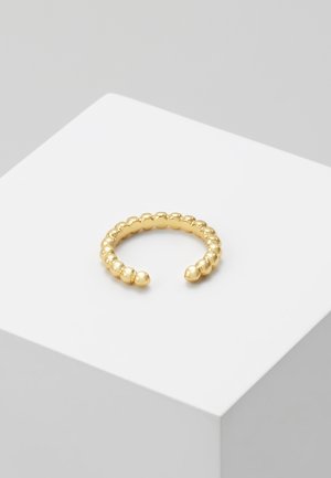 Bague ouverte en or avec un design perlé exposée sur un piédestal carré blanc sur fond gris.