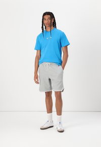 Modra bombažna majica s malim logotipom, sivi kratki hlače z belimi obrobami in bele superge. Model ima dolge pletene lase. Preprost ozadje.