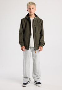 Green softshell jacket with a hood, zippered chest pocket, and elastic cuffs, paired with grey sweatpants and black sneakers.