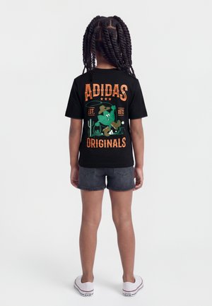 Bambino con capelli intrecciati che indossa una t-shirt nera Adidas Originals con un cartone animato di un cactus cowboy e testo arancione, abbinata a pantaloncini di denim e sneakers bianche.