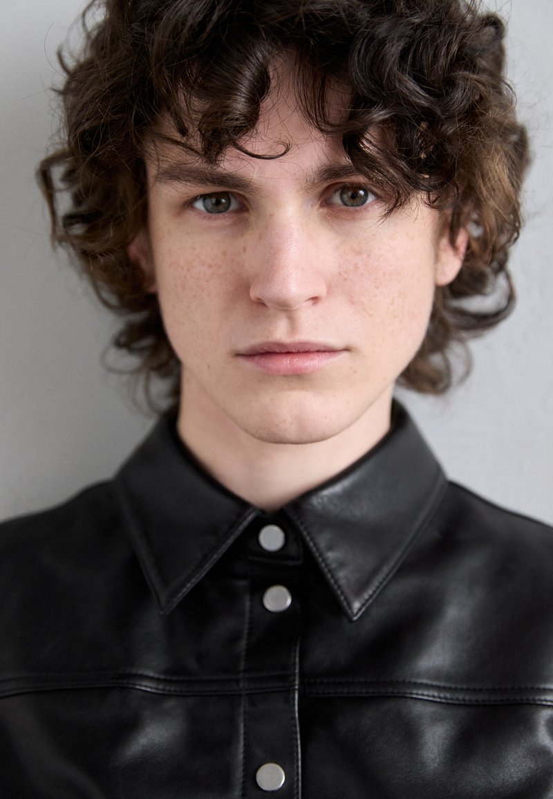 Chemise en cuir noir avec un col pointu, des boutons pression et une texture lisse, présentant des détails de couture visibles et une coupe ajustée.