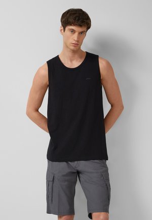 Mężczyzna w czarnej koszulce bez rękawów i szarych spodenkach typu cargo stoi na tle jasnoszarej ściany z rękami za plecami.