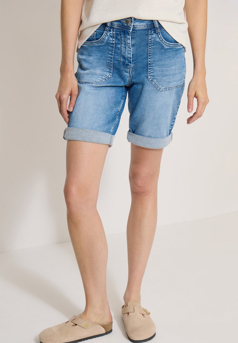 Personne portant un short en jean bleu roulé, des chaussures beige sans lacets et un haut de couleur claire, debout devant un fond uni.