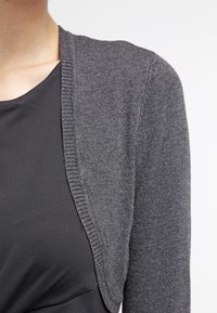 Grau gestrickte Strickjacke mit weicher Textur, offenem Schnitt und gerippten Kanten, kombiniert mit einem eng anliegenden schwarzen Oberteil.
