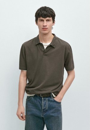 Jeune homme aux cheveux foncés portant un polo marron à manches courtes sur un t-shirt blanc et un jean bleu, une main dans la poche.