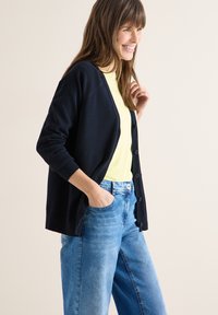 Femme souriante aux cheveux bruns portant un cardigan bleu marine sur une chemise jaune clair et un jean bleu, posant avec une main dans la poche sur un fond beige.