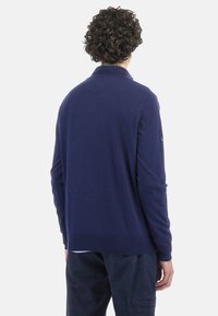 Navy blue sweater with a high collar, long sleeves, ribbed cuffs and hem, made of soft fabric. Features a logo on the left shoulder.