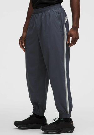 Uomo che indossa pantaloni da jogging grigio scuro con strisce laterali grigio chiaro e scarpe da ginnastica nere, in piedi davanti a uno sfondo semplice.