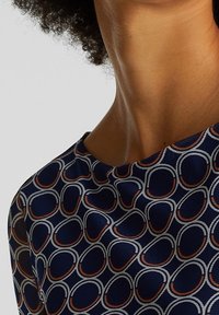 Mörkblå topp med rund halsringning, prydd med ett geometriskt mönster av överlappande cirklar i vitt och rostiga accenter. Texturerad tyg.