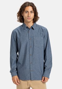 Blue long-sleeve button-up shirt made of textured fabric, featuring a chest pocket and brown buttons, worn with beige pants.