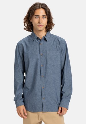 Blue long-sleeve button-up shirt made of textured fabric, featuring a chest pocket and brown buttons, worn with beige pants.