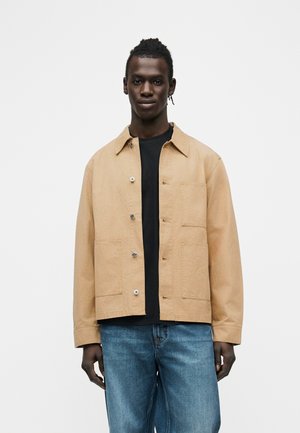 Mand stående iført beige knappet jakke over sort skjorte og blå jeans mod en ensfarvet hvid baggrund.