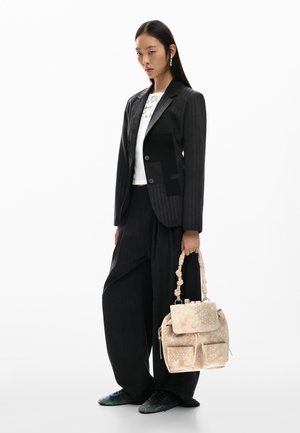 Vrouw in een zwart pantalonpak met krijtstreep en wijde broek, wit shirt, die een beige handtas met patroon en een geknoopte handgreep vasthoudt, staand tegen een witte achtergrond.