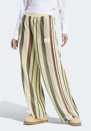 Labave pletene hlače s vertikalnim prugama u zelenoj, smeđoj, bež i sivoj boji, nošene s bež cipelama i bijelom bluzom dugih rukava.