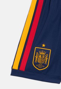 Μπλε ναυτικό αθλητικό σορτς με κόκκινες και κίτρινες πλευρικές ρίγες. Φέρει χρυσό έμβλημα με κορόνα και κείμενο "RFEF 1909" πάνω από ένα σχέδιο ασπίδας.