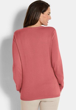 Donna sorridente, di spalle, che indossa un maglione a maniche lunghe di rosa polveroso e pantaloni beige chiaro su uno sfondo semplice e chiaro.