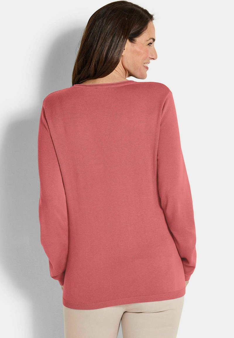Frau, die lächelt und mit dem Rücken zum Betrachter steht, trägt einen langärmligen, staublila Pullover und helle beige Hosen vor einem schlichten, hellen Hintergrund.