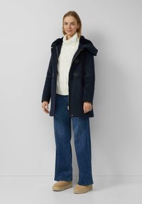 Marineblaue Parka mit Kapuze, ausgestattet mit Knebelknöpfen und Reißverschluss. Getragen über einem weißen, kabelgestrickten Pullover und ausgestellten Jeans.