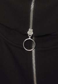 Giaccone nero con zip frontale dotata di una linguetta circolare, texture a coste nel colletto e nei polsini, e tessuto liscio.