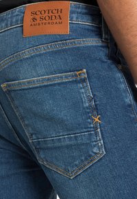 Tmavě modré džíny z denimu s hnědou koženou nášivkou označenou "SCOTCH & SODA AMSTERDAM," s oranžovým prošíváním a detailem na zadní kapse.