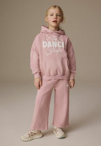 Rosa hoodie med vit "DANCE Studio" grafik, tillsammans med matchande vida rosa byxor. Bärs med ljusa sneakers. Mjuk tygtextur.