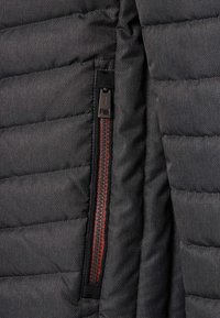 Sezione di giacca a vento nera con texture trapuntata, zipper con accenti rossi e cuciture rinforzate delle tasche. Il materiale sembra resistente e liscio.