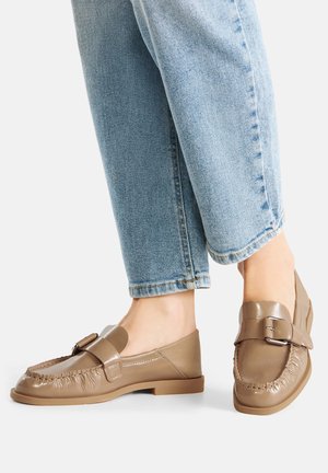 Mocasines de cuero marrón con detalle de flecos texturizados, suela plana y adorno de hebilla, combinados con vaqueros azul claro con puños.