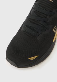 Μαύρο αθλητικό παπούτσι με επάνω μέρος από διαπνέον διχτυωτό υλικό, υφή στην επιφάνεια, μαύρα κορδ όνια και χρυσές λεπτομέρειες στο πλάι και στη μύτη.