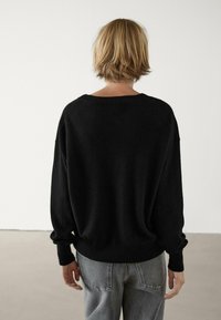 Personne avec les cheveux blonds courts portant un pull noir ample et un jean gris, se tenant face à un mur blanc uni.