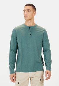 Langærmet teal henley skjorte med tre sorte knapper ved halsen, der har en glat overflade og lette sideslidser for komfort.