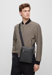 Czarna skórzana torba na ramię o teksturowanym wykończeniu, z zamknięciem na zamek błyskawiczny i regulowanym paskiem, zestawiona z kurtką bomberką w kratkę.