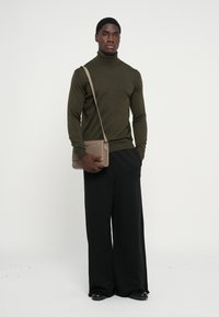Maglione a collo alto verde oliva, pantaloni neri a gamba larga, in mano una borsa a tracolla beige con zip, texture liscia, design minimalista.