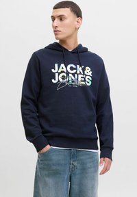 Marineblaue Kapuzenjacke mit grafischem Text "JACK & JONES Essentials" auf der Vorderseite, ausgestattet mit einer verstellbaren Kapuze und gerippten Bündchen. Kombiniert mit hellblauen Jeans.