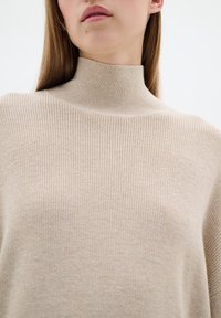Beige geribbelde turtleneck trui met een zachte textuur, voorzien van een aansluitende kraag en een losse silhouet, benadrukt een minimalistisch ontwerp.