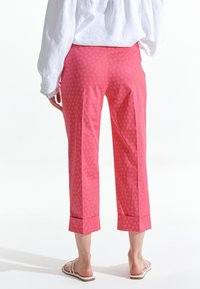 Bakifrån av en person som bär ljusrosa cropped byxor med gula stjärnmönster, vit långärmad tröja och vita sandaler mot vit bakgrund.