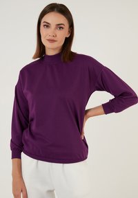 Felpa viola con colletto alto, maniche lunghe e polsini a coste. Texture morbida e vestibilità ampia, con tasche laterali e orlo curvato.