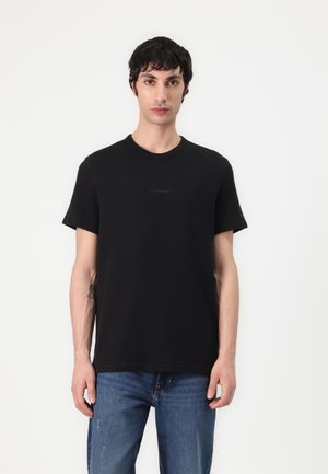Black t-shirt made of cotton, featuring a round neck, short sleeves, and a subtle logo on the front. Worn with blue jeans.