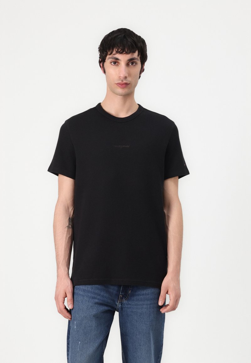 Camiseta negra de algodón, con cuello redondo, mangas cortas y un logo sutil en la parte delantera. Llevada con jeans azules.