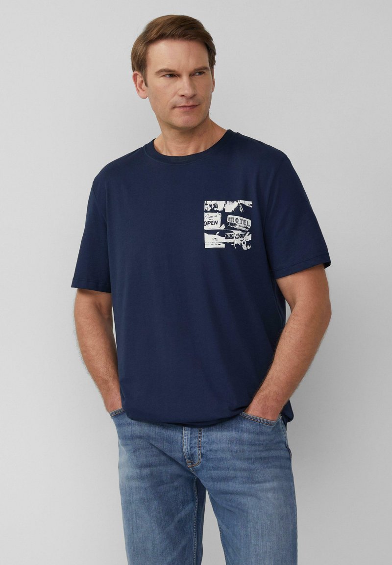 Marineblauwe katoenen T-shirt met korte mouwen, met een witte grafische zakontwerp van vintage borden, gecombineerd met lichtblauwe jeans.