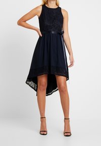 Robe de navire avec un corsage en dentelle, un ourlet asymétrique et une ceinture en satin. La jupe est ornée d'une bordure en dentelle texturée en bas.